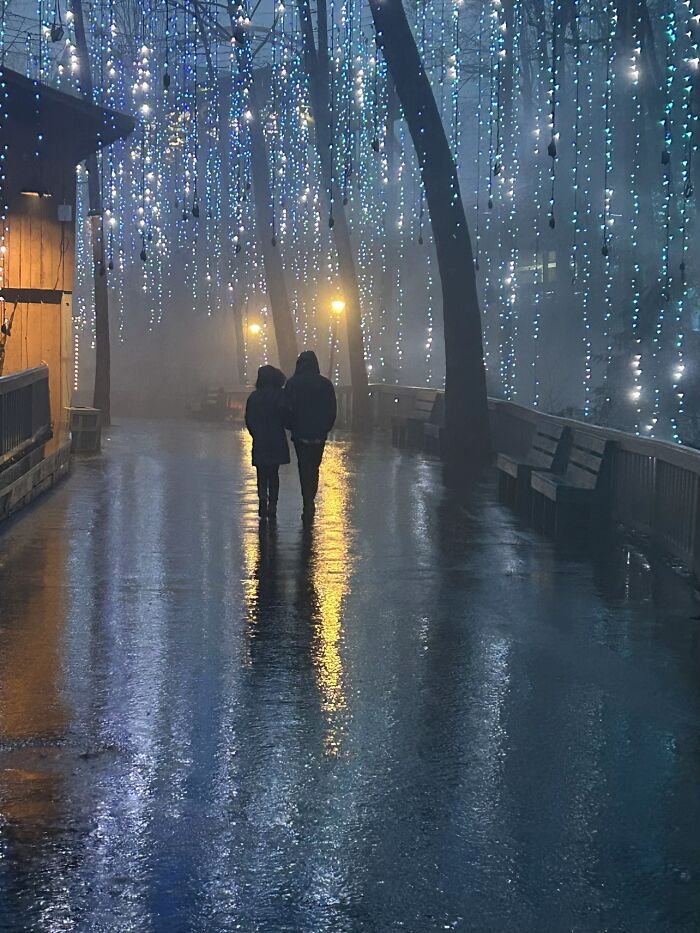 Two strangers walk in a misty alley adorned with glowing lights, capturing a wholesome photo trend.