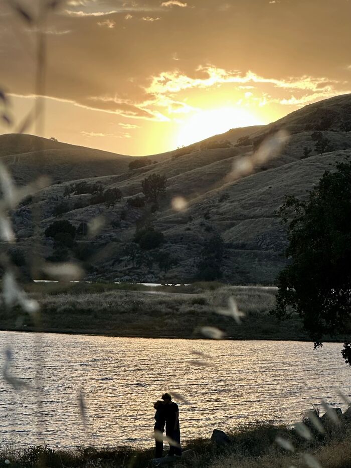 A wholesome photo of two strangers by a lake at sunset, resembling a painting with hills in the background.