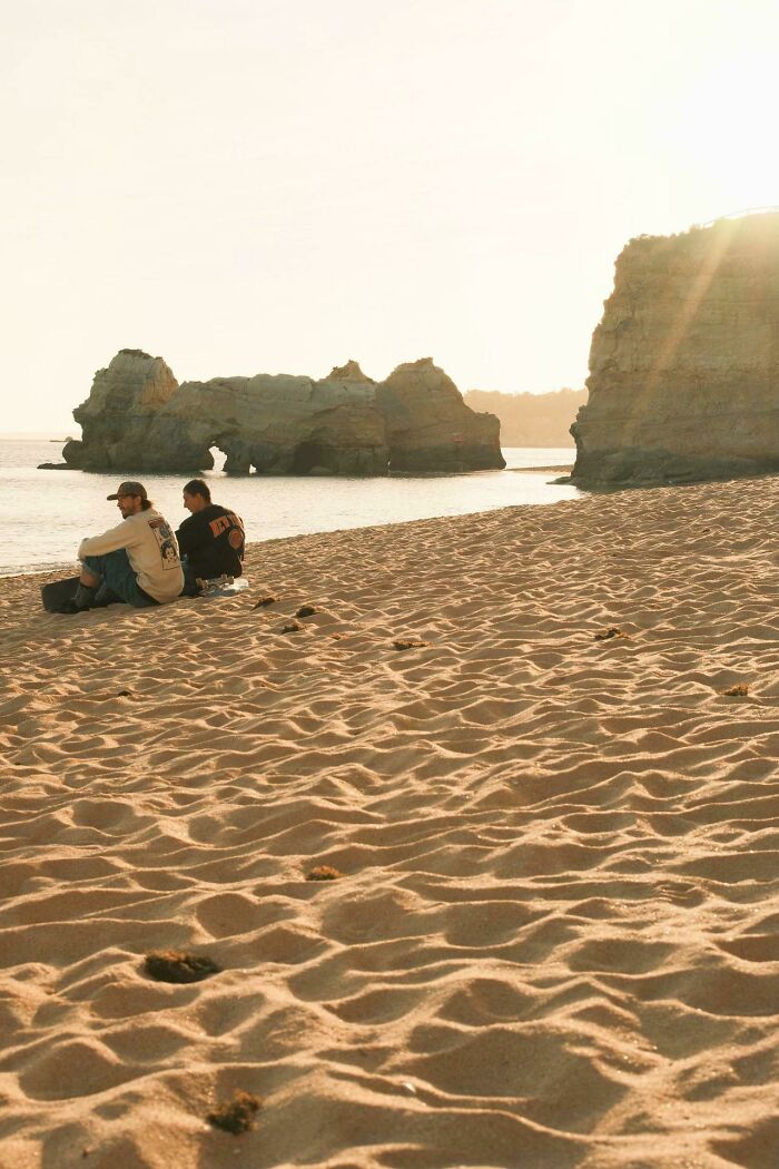 Wholesome photo trend: two strangers sitting on a sandy beach, enjoying the painting-like sunset with sea arches.