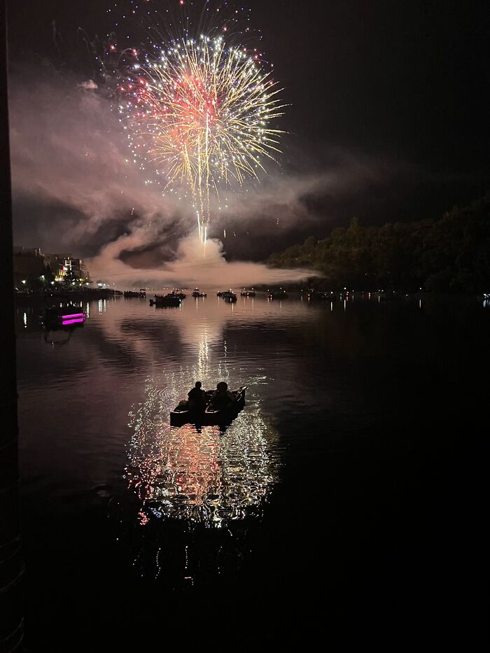 People in a boat watch colorful fireworks reflected on a dark lake, part of a wholesome photo trend.
