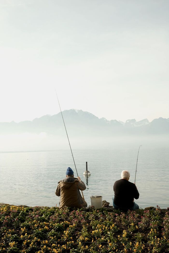 A wholesome photo trend of two men fishing by a serene lake with misty mountains in the distance.