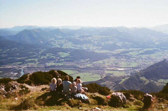 A group of people sit on a mountain peak, gazing at a vast landscape below, embodying a wholesome photo trend.