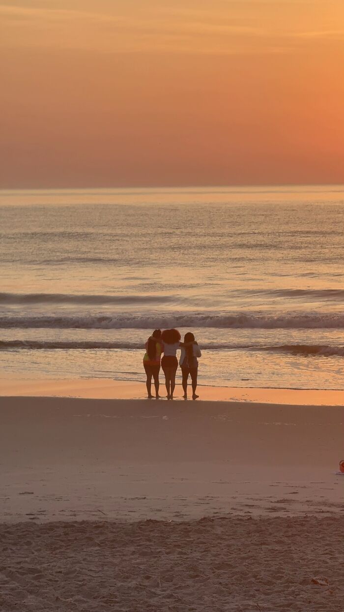 A wholesome photo trend featuring three friends standing together on a beach, silhouetted against an orange sunset.
