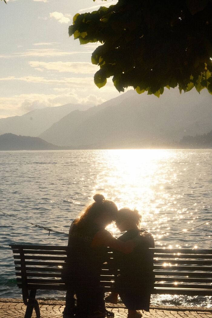 A wholesome photo trend of two people embracing on a bench by a shimmering lake with mountains in the background.