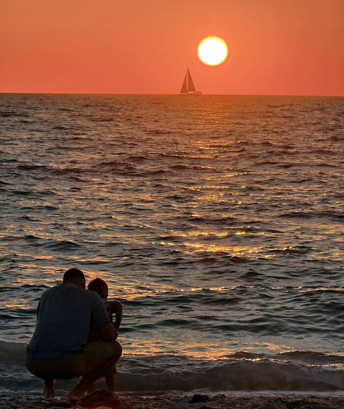 A wholesome photo trend showing a father and child watching a beautiful ocean sunset with a sailboat.