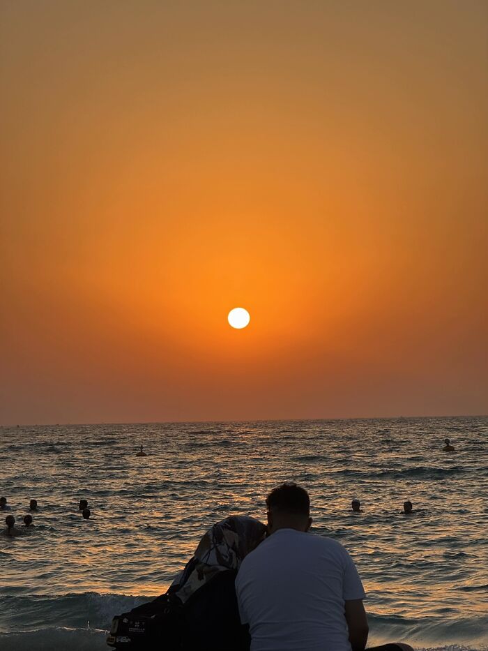 A couple watching the sunset over the ocean, like a painting, embodying the wholesome photo trend between strangers.