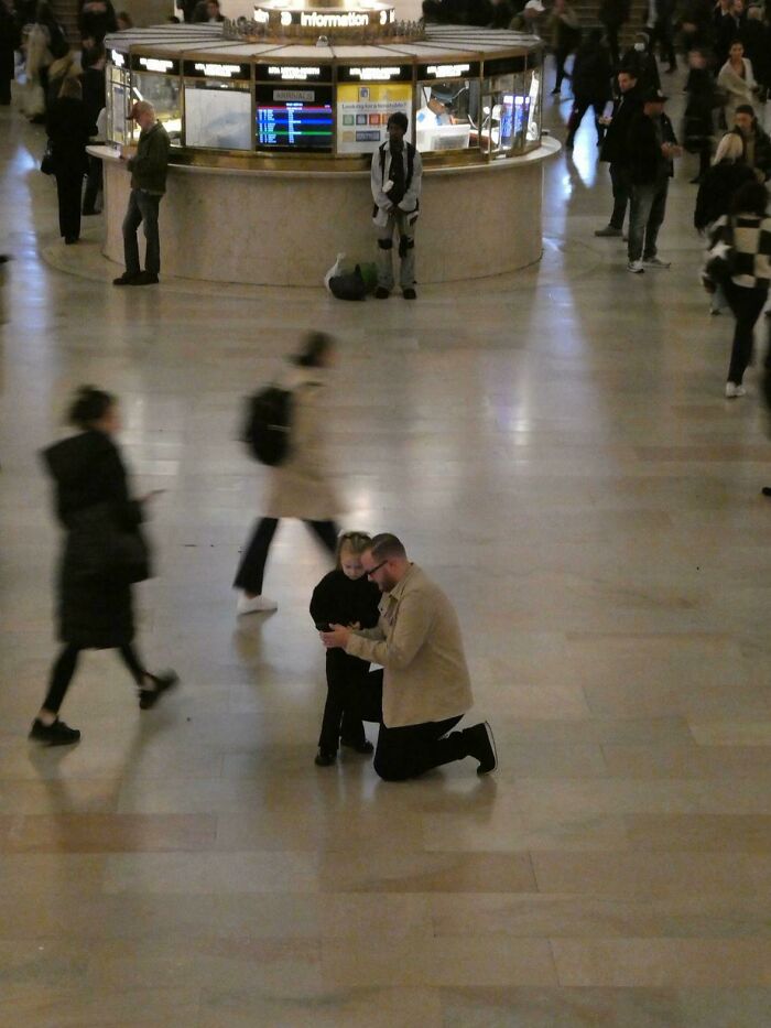 A father kneels to interact with his child in a busy station, a painting-like wholesome photo moment.