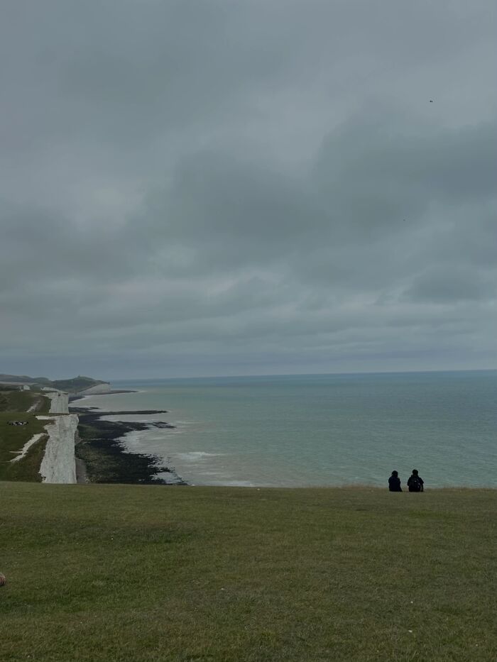 Two people sitting on a grassy cliff overlooking the ocean, embodying the 'Like A Painting' photo trend.