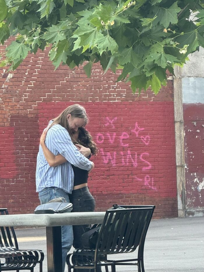 A wholesome photo of a couple hugging in front of a red brick wall with graffiti, part of the sweet photo trend.
