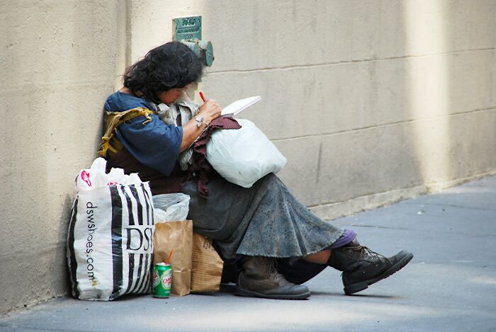 Homeless woman writing in notebook on street with bags and soda nearby