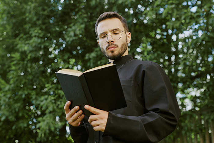 Man in black clerical clothing reading a book outdoors