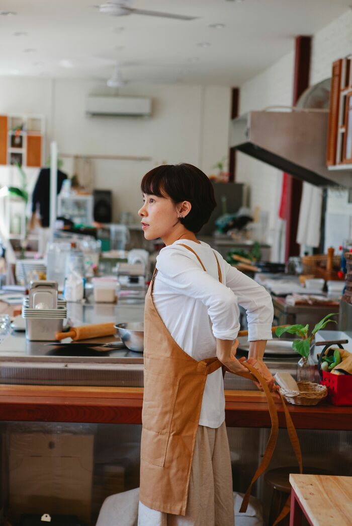 A woman in an apron in a kitchen, looking pensive, perhaps after hilariously misreading social cues and making things awkward.