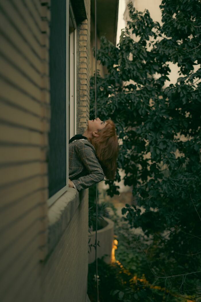 Woman leaning backward from a window looking up surrounded by trees