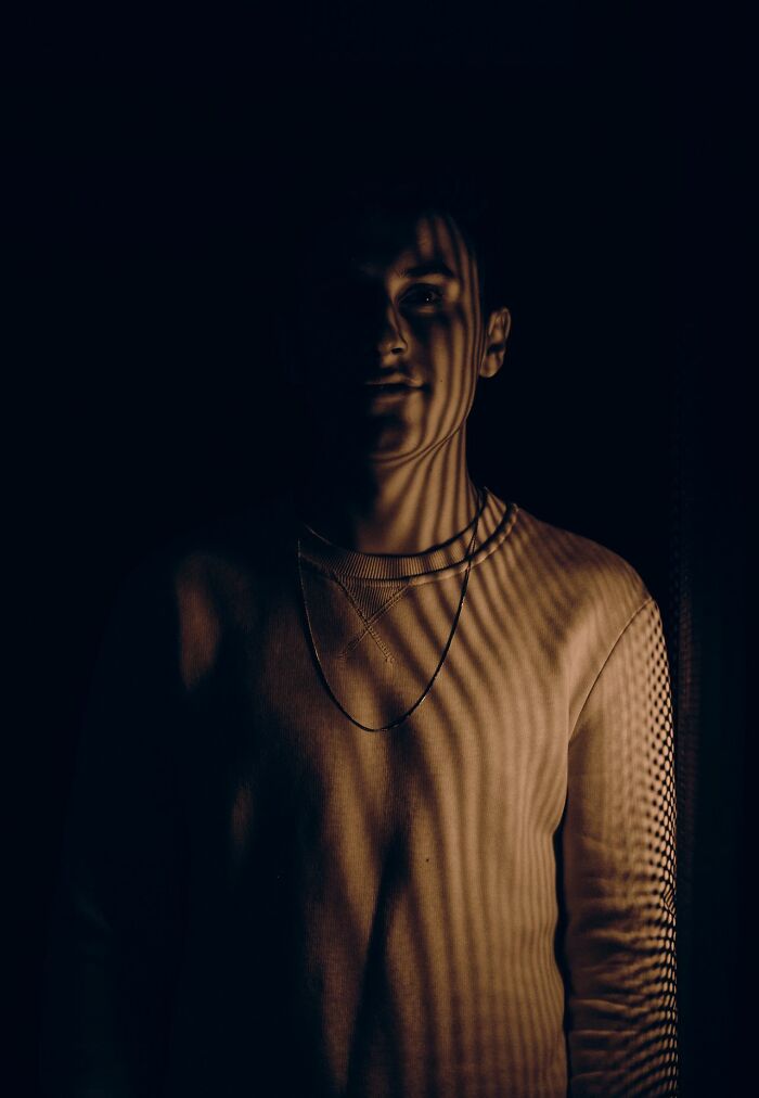 A man with striped shadows on his face and chest, wearing a light-colored sweater and necklace, illustrating experiences people can't explain.