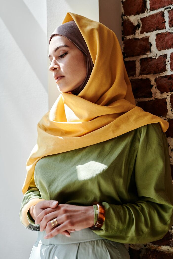 A woman in a yellow hijab looks down with a pensive expression, considering the experiences of polygamous families.