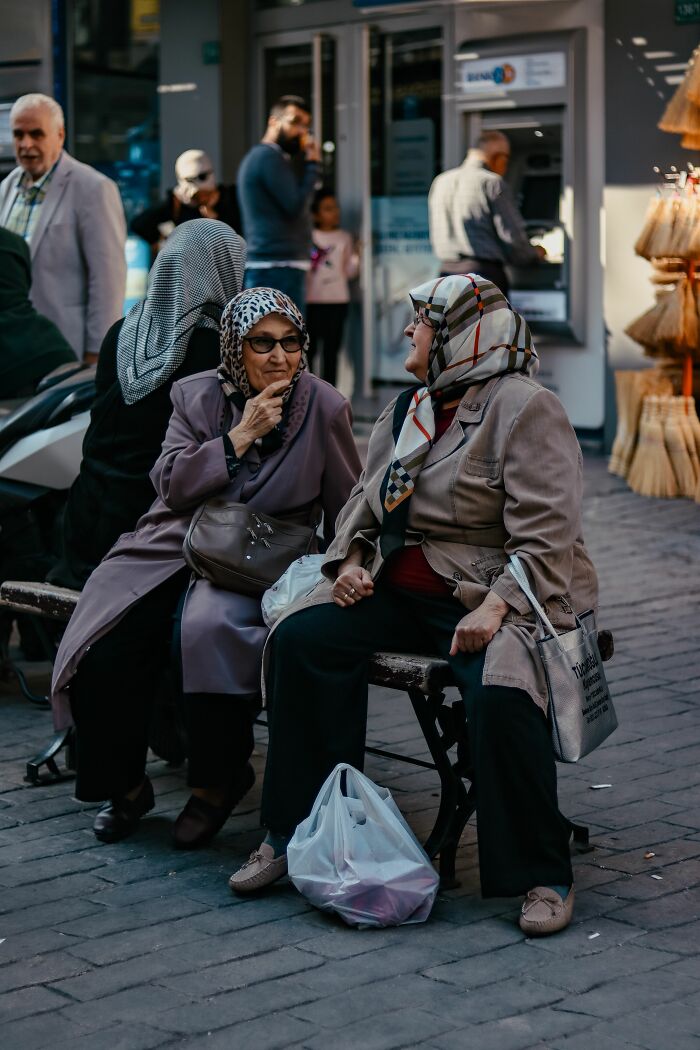 Two elderly women in headscarves sitting and conversing on a street bench, depicting interactions among locals and foreigners.