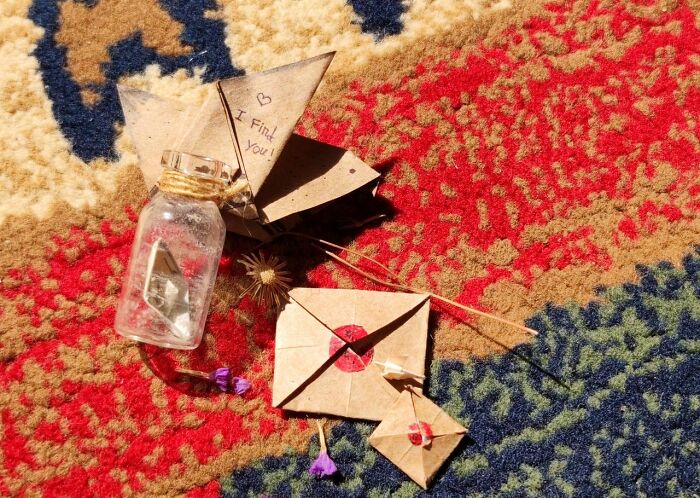 A small glass bottle, paper origami with heart, and tiny paper envelopes arranged on a colorful patterned rug, symbolizing experiences people can't explain.