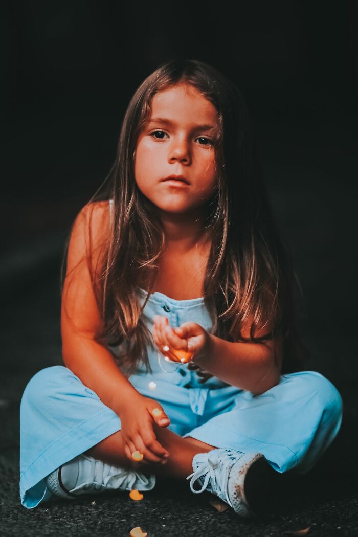 A young girl with long hair sits cross-legged, holding small objects, with an unexplainable look.