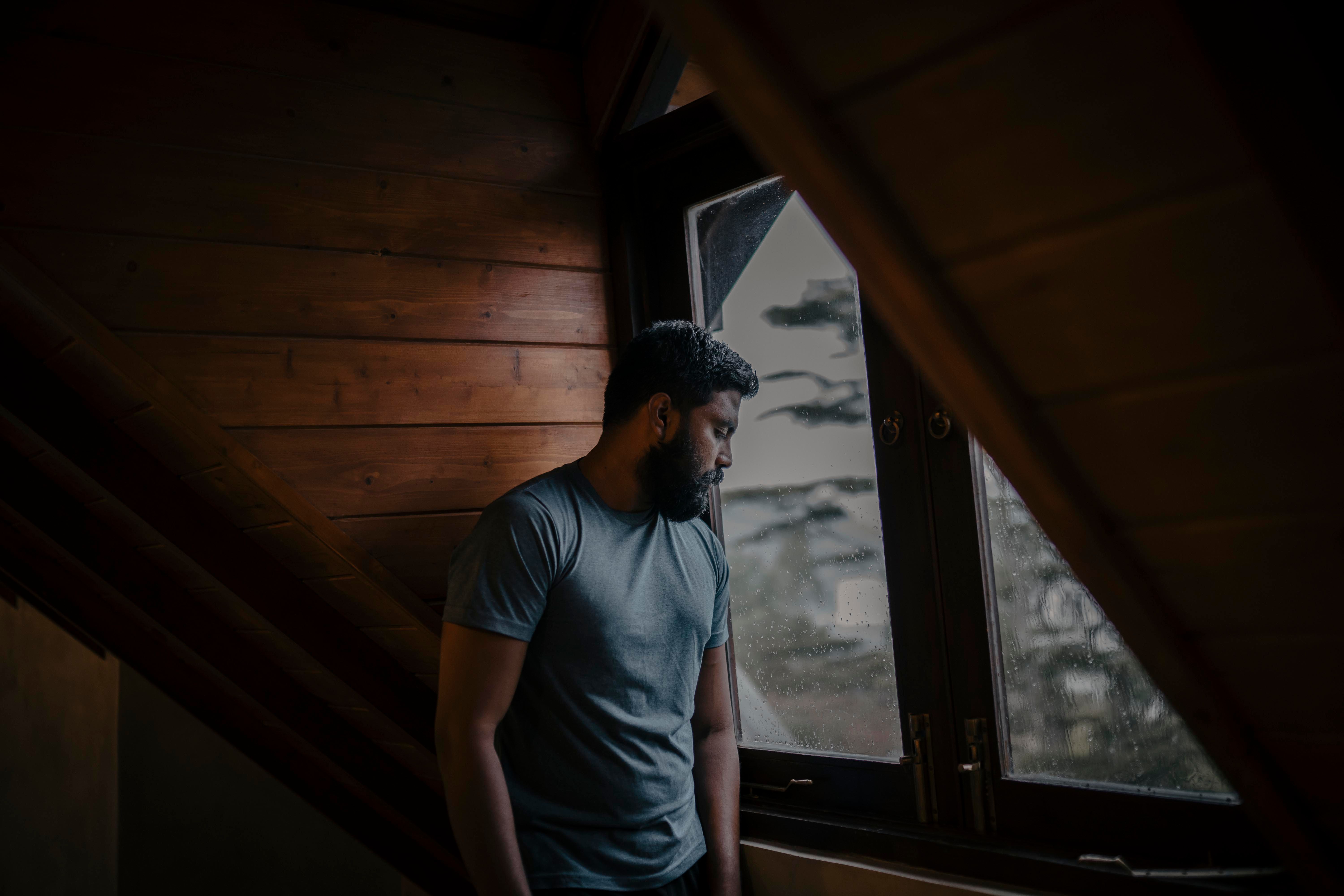 Man looking sadly out a rainy window in a wooden room Man looking sadly out a rainy window in a wooden room