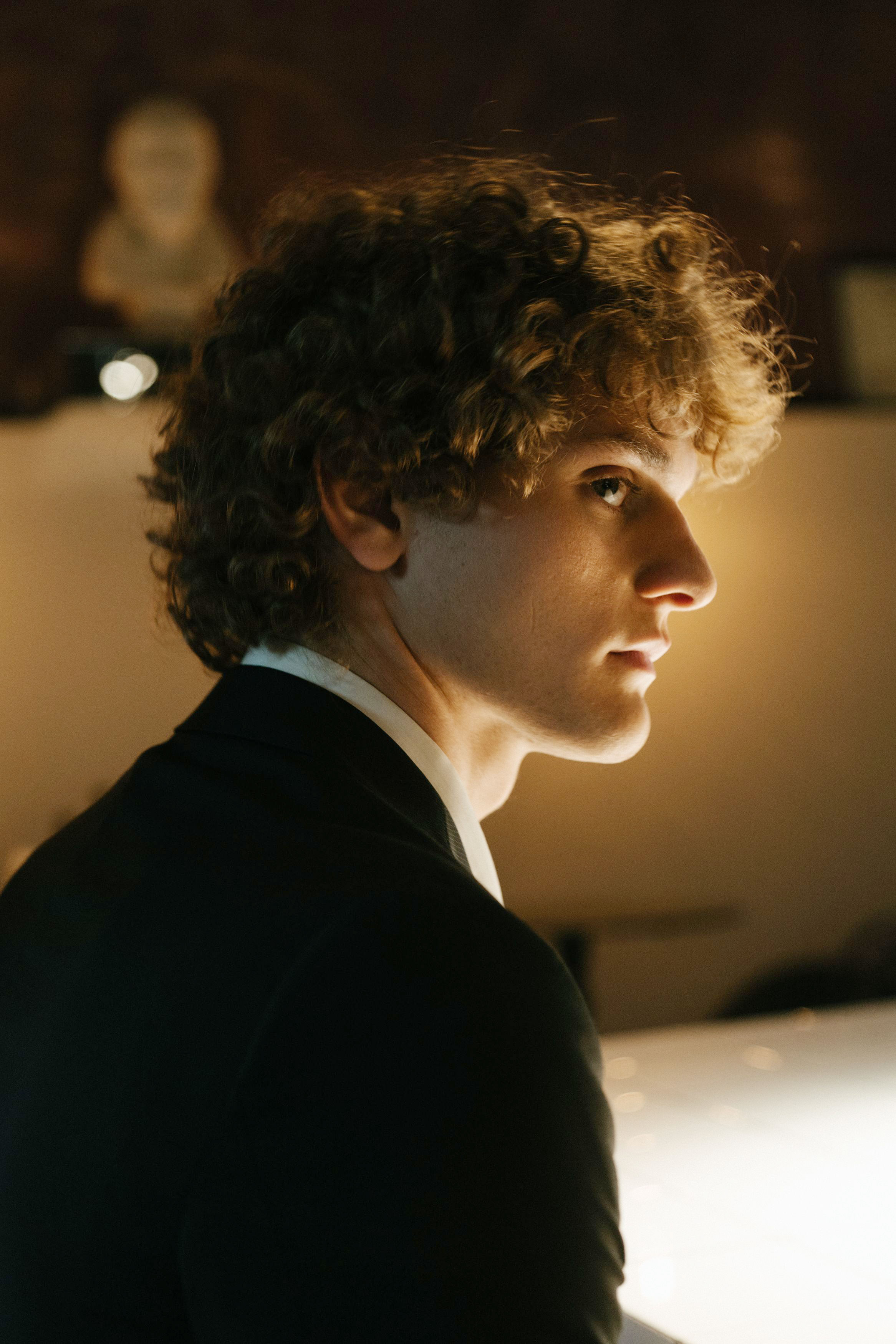 Man with curly hair, wearing a suit, looks away from the camera, perhaps during a rehearsal dinner spectacle. Man with curly hair, wearing a suit, looks away from the camera, perhaps during a rehearsal dinner spectacle.
