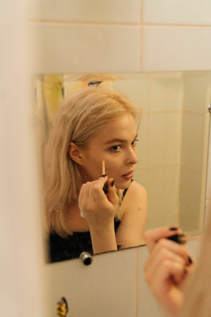 A blonde woman in a mirror applies makeup, highlighting a beauty routine that might be a turn off for some.