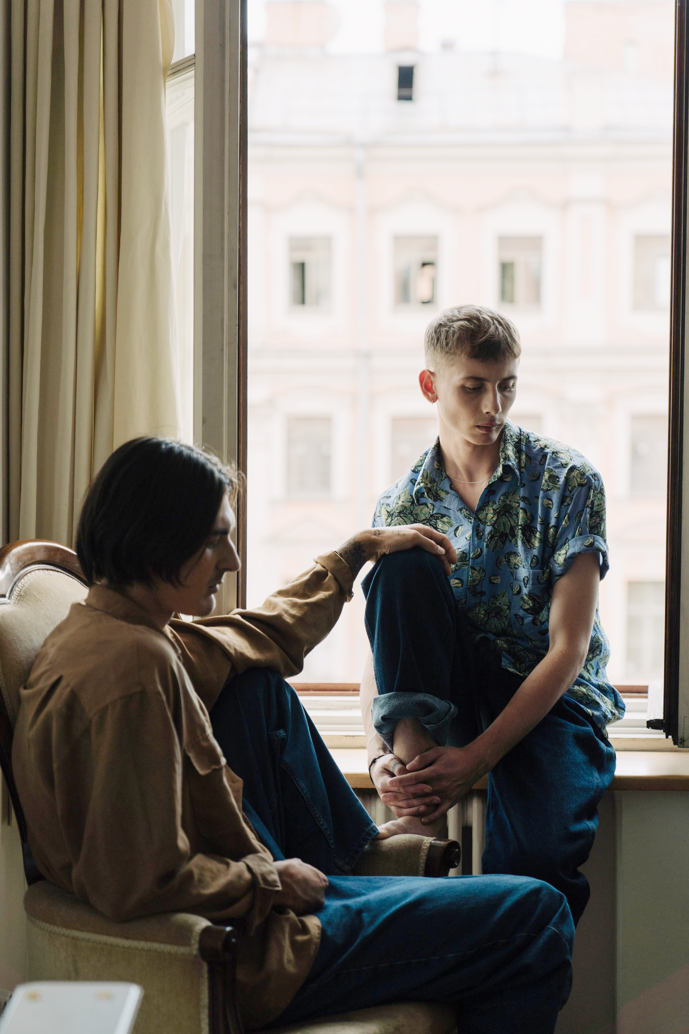 A man sitting on a window sill looks at another man sitting in a chair, discovering a double life.