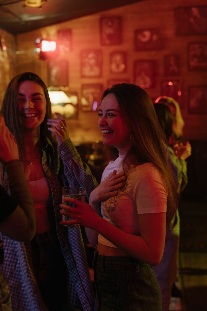 Two women laughing in a dimly lit bar, sharing an unbelievable coincidence they experienced.