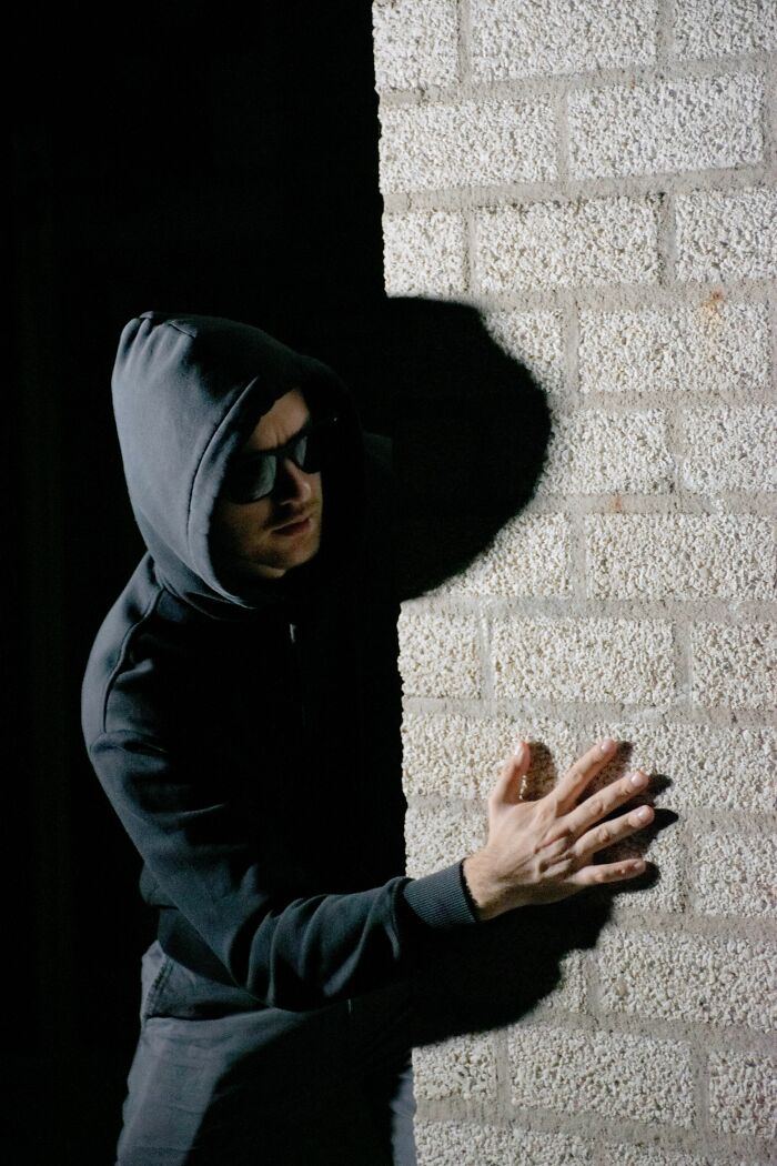 A person in a dark hoodie and sunglasses peeking from behind a brick wall, symbolizing hidden true selves.