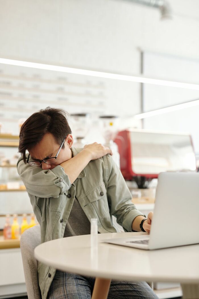 A man in glasses coughing into his elbow while sitting at a table with a laptop, an unserious ER visit scenario.