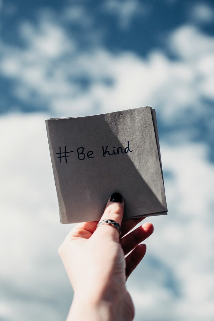 A hand holding a Be Kind note against a cloudy sky, a hint showing a person is secretly very smart.