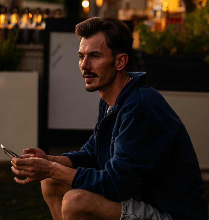 A thoughtful man, visiting the US, looks to the side while holding a phone in an outdoor setting.