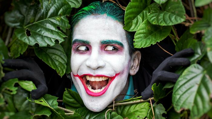 Close-up of movie villain with green hair and white face paint peeking through leaves