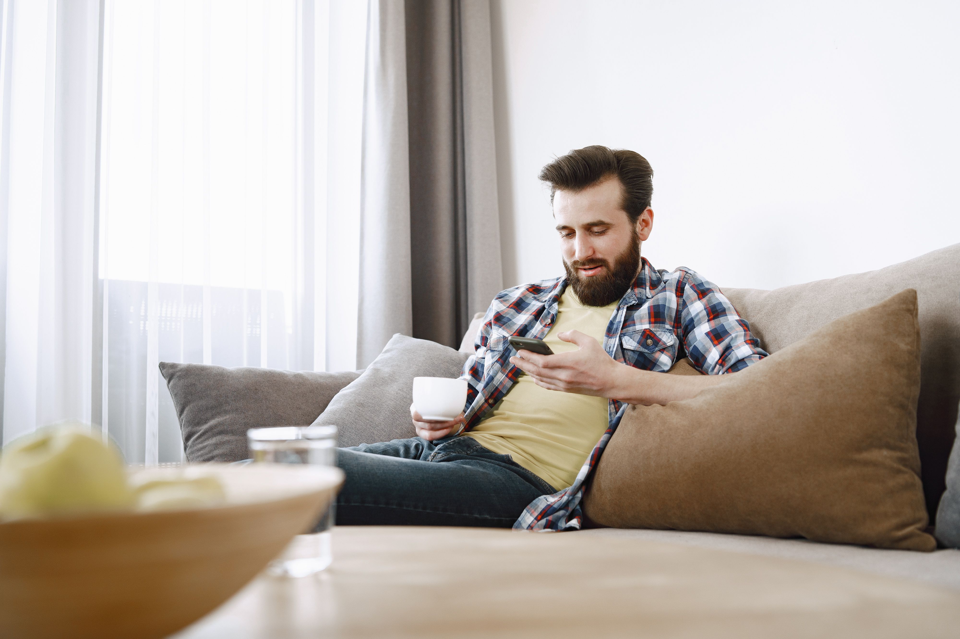 Unemployed man looking at phone on couch with coffee cup Unemployed man looking at phone on couch with coffee cup
