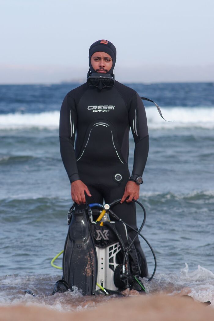 Scuba diver in wetsuit standing on shore with diving gear and fins by ocean waves