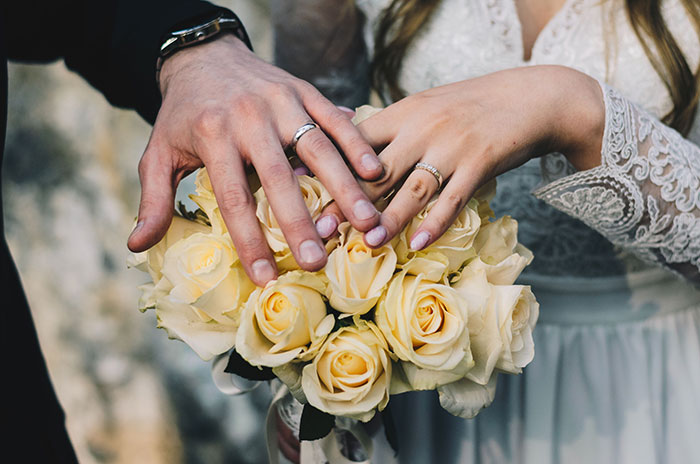 Bride and groom's hands with wedding rings resting on a bouquet of white roses.