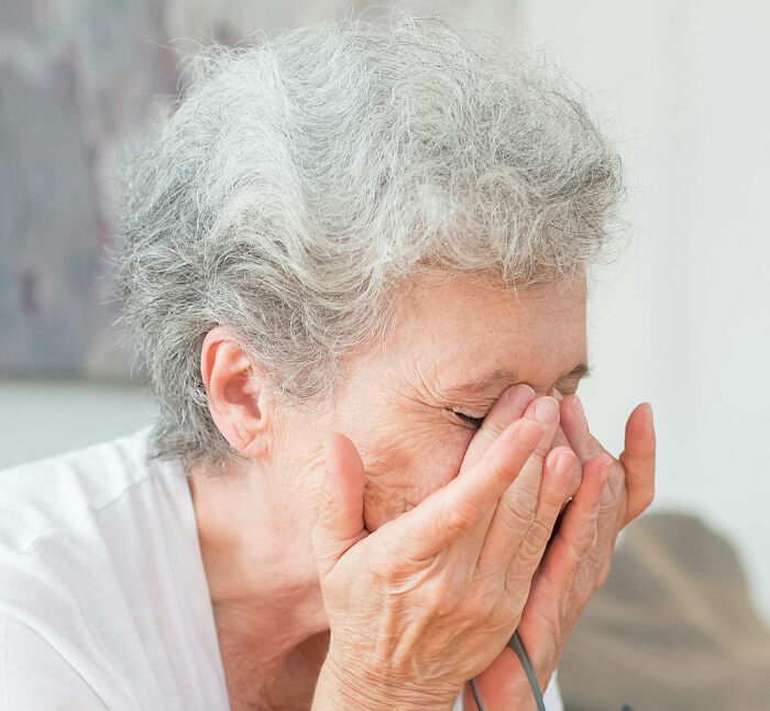 An 80-year-old woman with gray hair covering her face with her hands, looking distressed, after encountering a Male Karen.