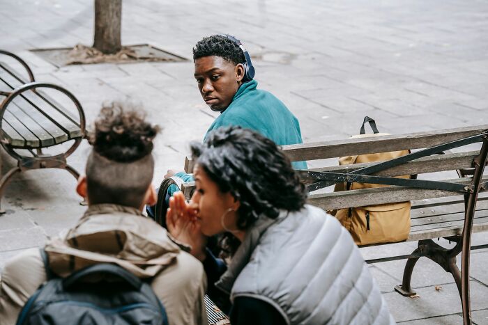 Young man appearing anxious while sitting on a bench with two people whispering nearby, anxiety thought patterns