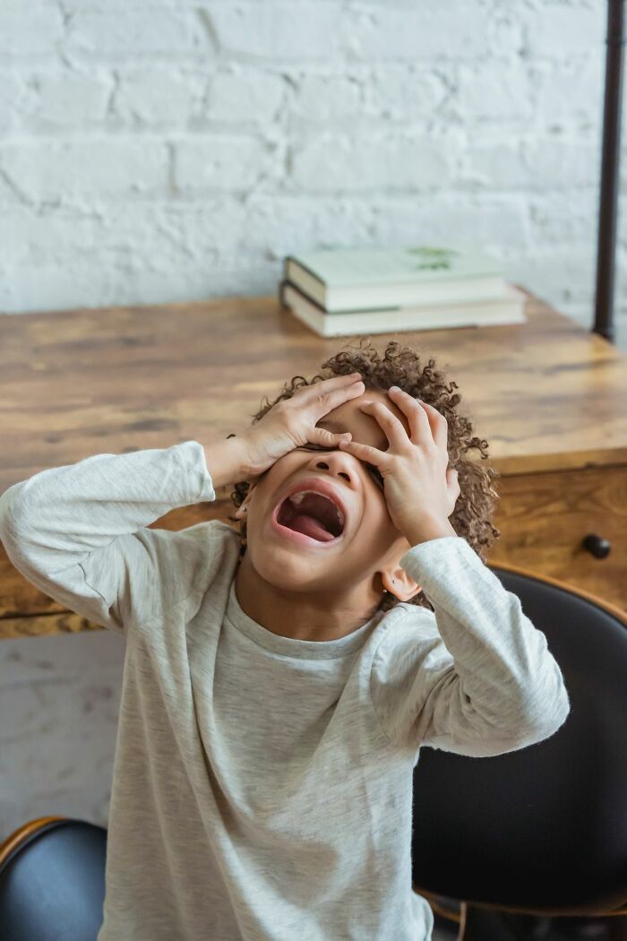 A child with curly hair screams, holding their head in frustration or pain at a desk.