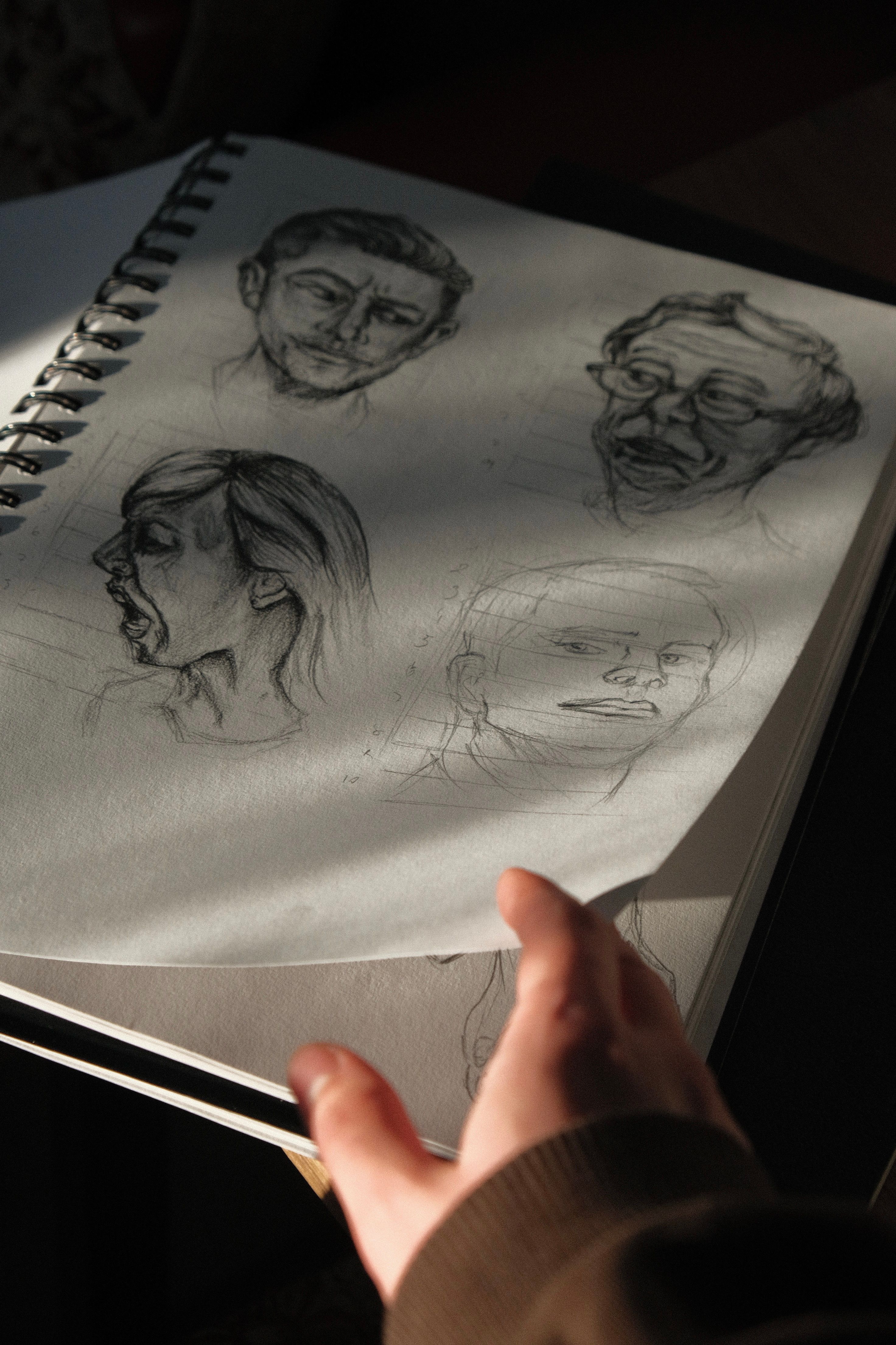 A hand flips through a sketchbook showing various facial expressions, part of a husband living a double life.