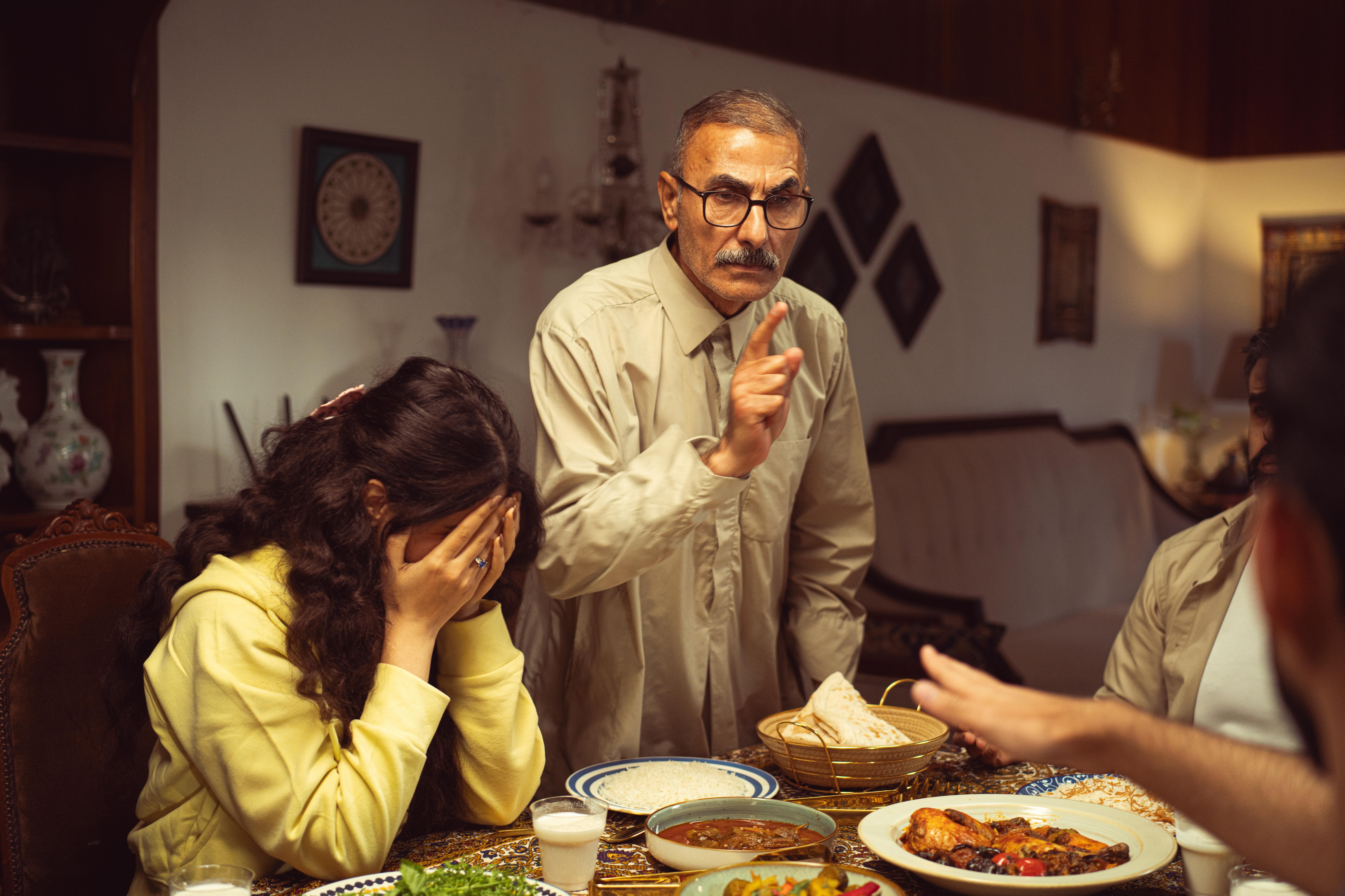 An older man with glasses points an accusing finger at a woman covering her face at a dinner table during a family vacation.