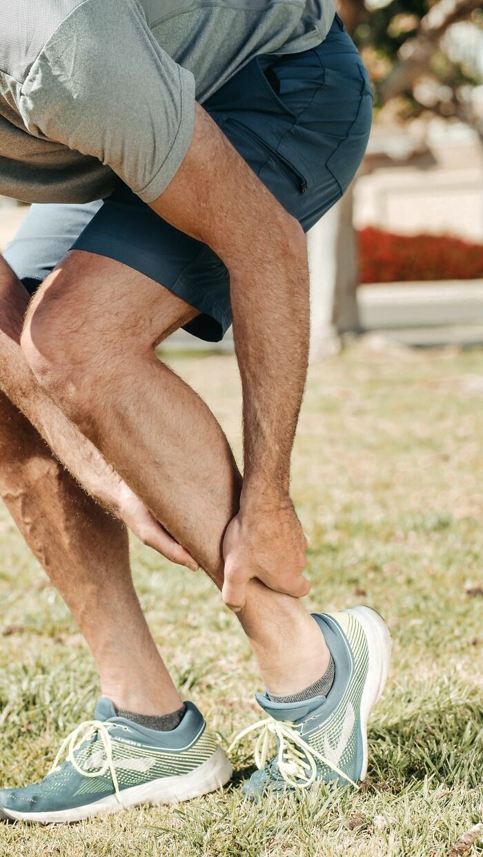 A man grasps his calf in pain while standing outdoors, showing physical discomfort.