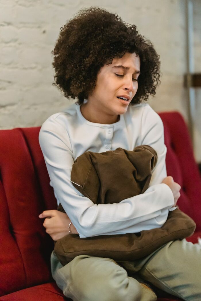 A woman with curly hair, eyes closed, clutches a pillow on a red couch, enduring internal pain.
