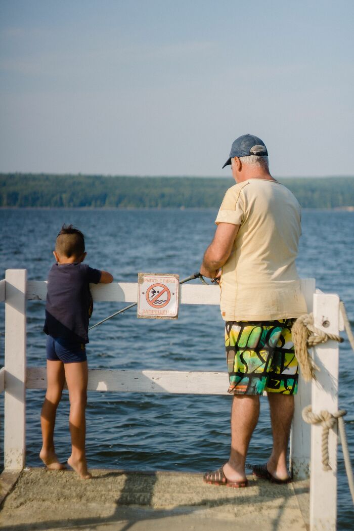 A man and a child hide their true selves on a pier, looking out at the water with a no fishing sign.