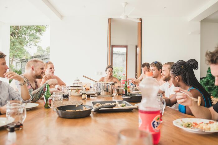 A diverse group of locals and foreigners shares a meal at a long table, highlighting potential cultural misunderstandings.