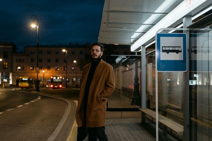 Man in brown coat waiting at a night bus stop under street lamps