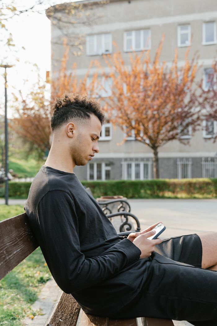 Young man sitting on bench, absorbed in phone after learning about cheating