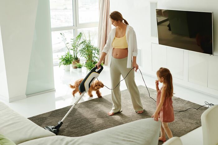 A pregnant woman vacuuming with a child and a dog nearby, showcasing human habits that can confuse dogs.