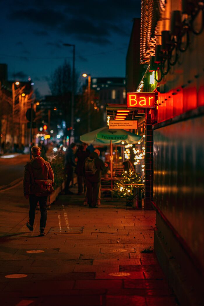 A street scene at night, with a person walking past a bar with a neon sign and outdoor seating. This is a wild coincidence.