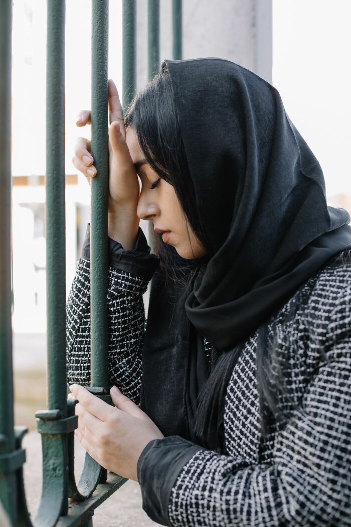 A woman in a hijab leans her head against a green fence, conveying the struggles of polygamous families.