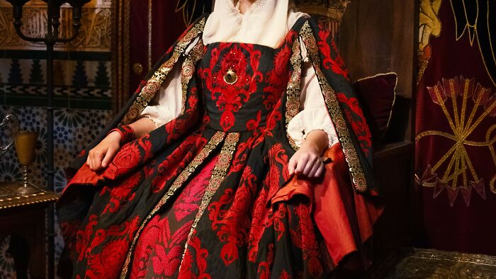 A regal character in an elaborate black and red brocade gown with gold trim, inviting viewers to identify TV series from outfits.
